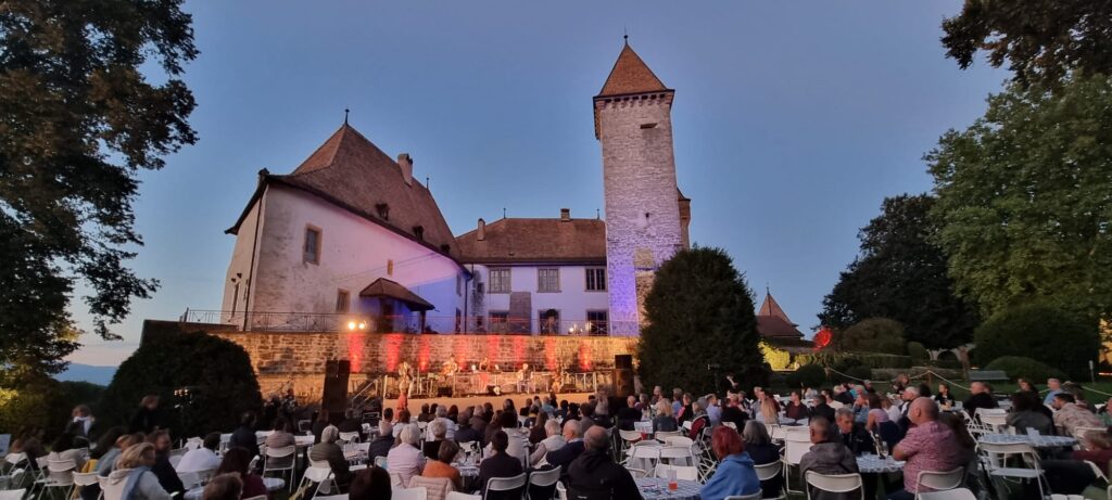 Photo prise depuis les jardins présentant le public venu en nombre écouté un groupe de musique, par un beau soir d'été